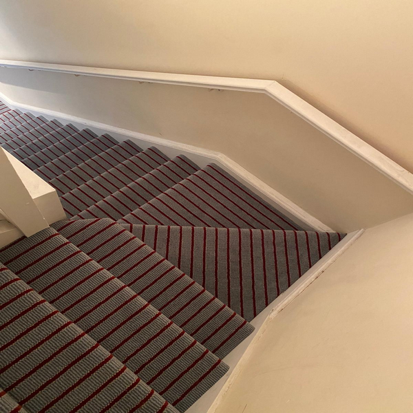 A carpeted staircase featuring a striped grey and red pattern, with beige walls and a white handrail.