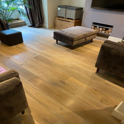 Living room with wooden floor, two brown armchairs, an ottoman, a TV on the wall, and a stack of firewood. A potted plant is near the window.