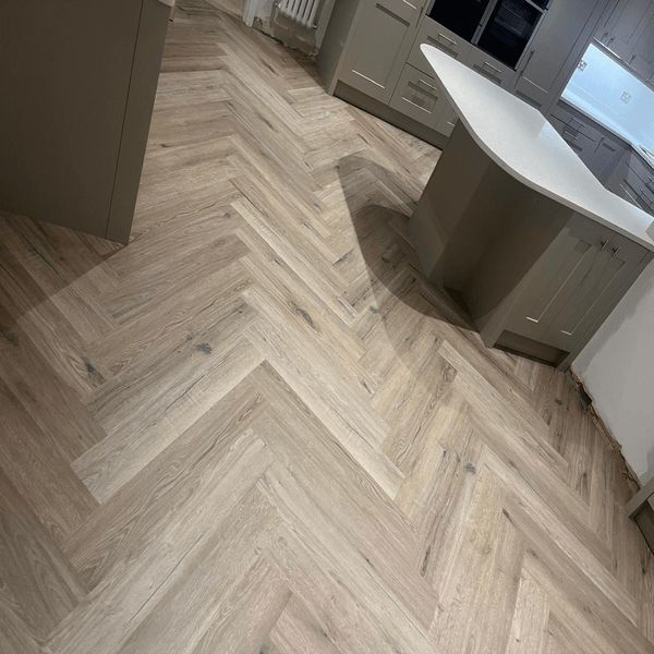 Herringbone-patterned wood floor in a modern kitchen with light gray cabinets and a curved white island. Luxury wood-effect LVT flooring installed in a Harrogate kitchen