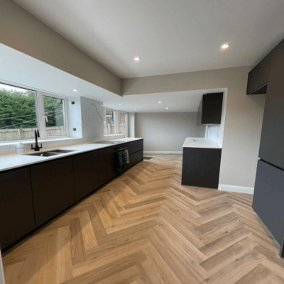 Modern kitchen with dark cabinets, white countertops, and herringbone wood flooring. Large windows provide natural light.