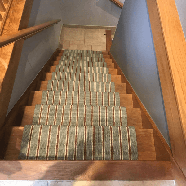 A wooden staircase with a striped carpet runner, featuring light brown and blue stripes, descends towards a tiled floor. The walls are painted in a light grey colour.